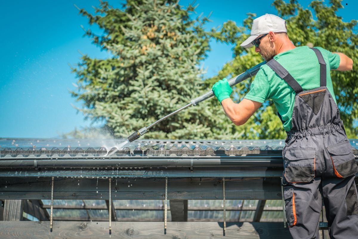 A person in overalls and gloves cleans a roof with a pressure washer, surrounded by greenery and a clear blue sky.