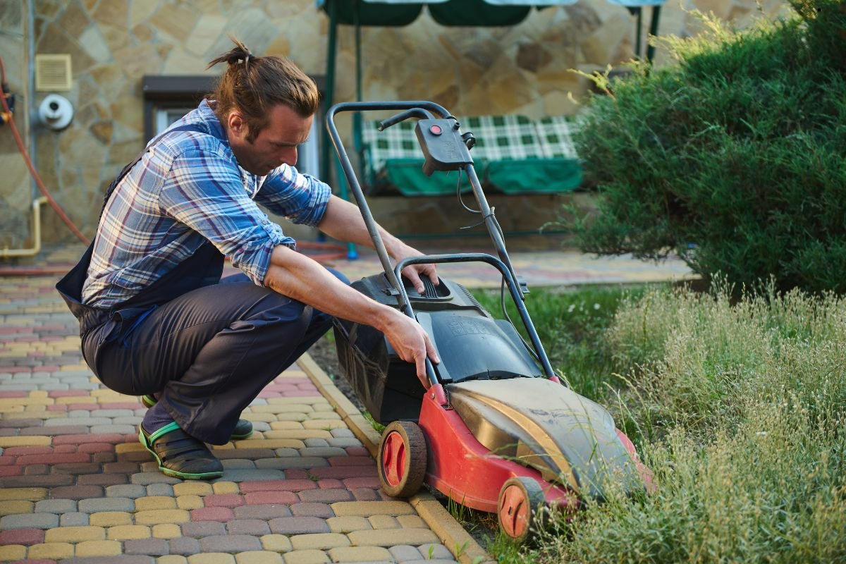 A person in plaid shirt crouches to adjust a red lawnmower on a paved pathway surrounded by overgrown grass and greenery.
